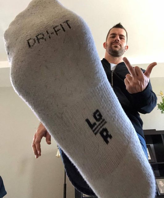 A man in a dark jacket and jeans shows his socked foot and gives the middle finger.