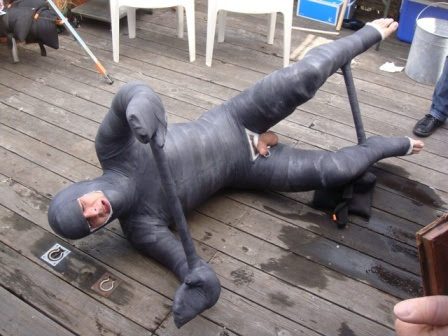 A man encased in a dark full body cast lies on a wooden deck with a cock ring visible.