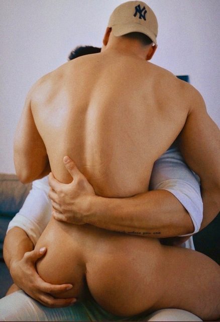 A muscular shirtless man wearing a baseball cap is held from behind by another man, showcasing his bare buttocks and broad back.