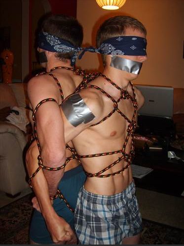 Two blindfolded men are tied together back-to-back with rope and gagged with duct tape indoors.