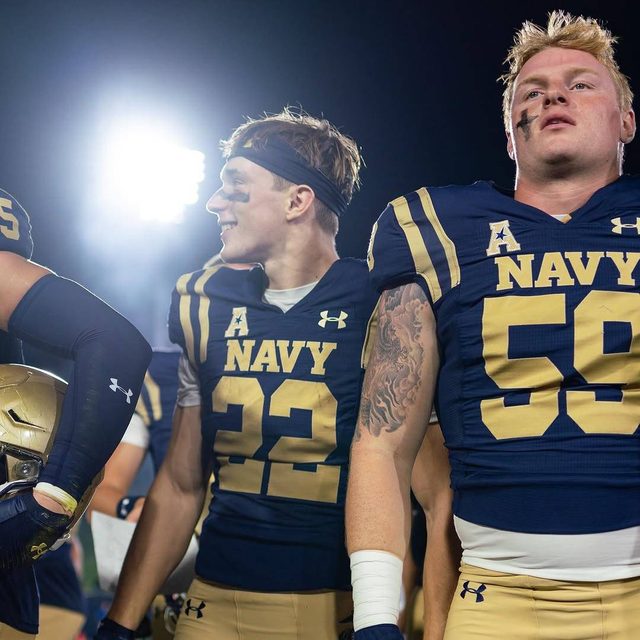 A group of athletic football players in full Navy uniforms stand together on the field under bright stadium lights at night.