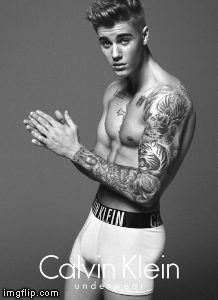 A tattooed, shirtless young man poses in white boxer briefs against a plain studio background in a black and white portrait.