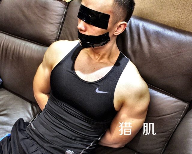 A muscular man in sportswear is blindfolded and gagged with tape while sitting on a couch.