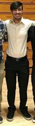 A smiling man stands in a group wearing a collared shirt, black pants, and sneakers.