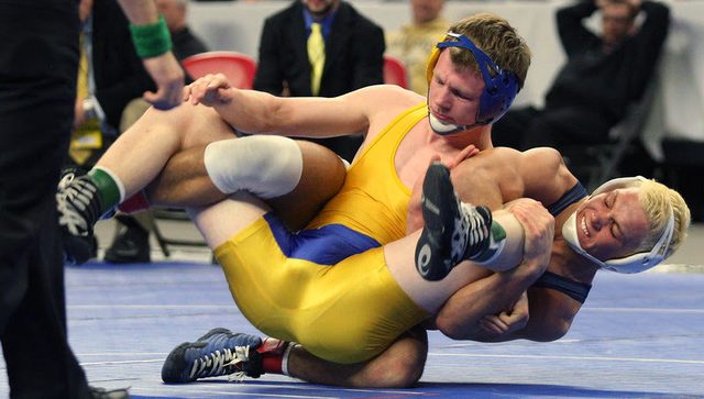 Two muscular men in wrestling singlets and headgear are engaged in a wrestling match on a blue mat.