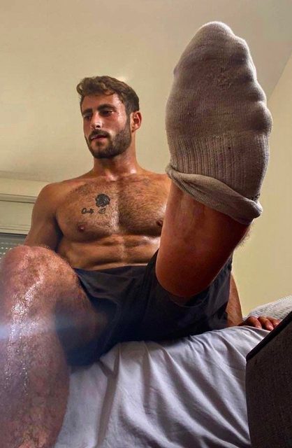 A sweaty, muscular man with a hairy chest sits on a bed and presents his socked foot to the camera.