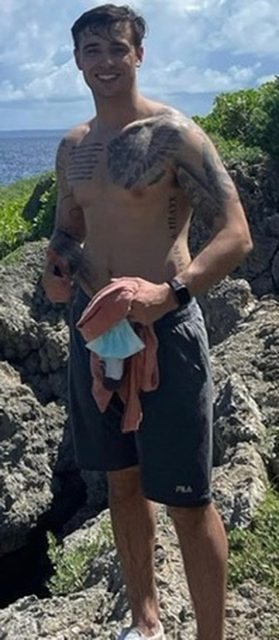 A smiling muscular man with tattoos stands shirtless outdoors on a rocky coast.