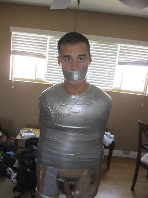 A man is restrained with tape around his body and his mouth gagged in a bedroom setting.