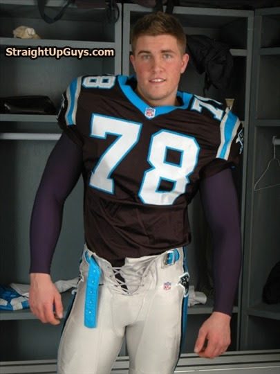 A young athletic man poses in a football uniform inside a locker room setting.