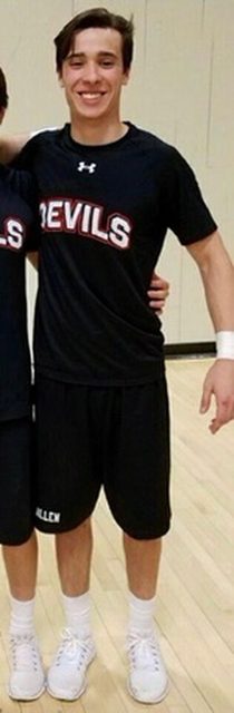 A smiling young man in a black t-shirt and shorts stands on a basketball court.