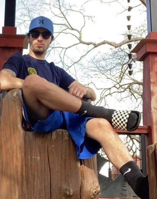 A man in a t-shirt, shorts, socks, and sandals sits outdoors on a wooden structure.
