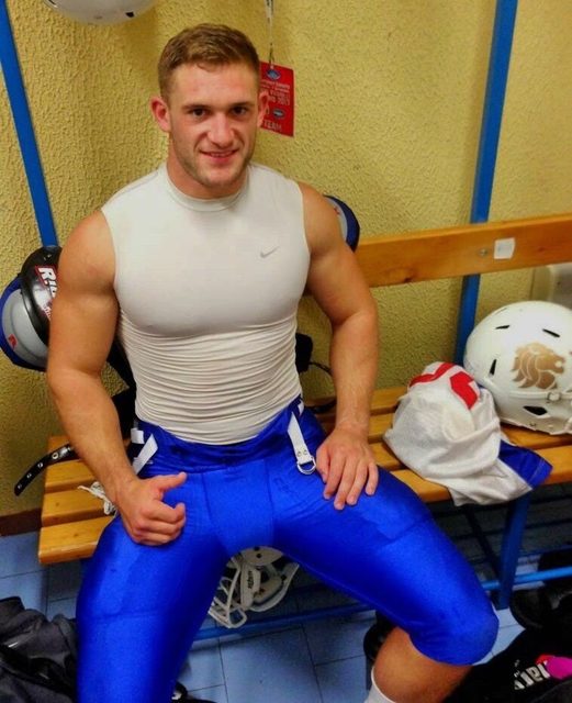 A muscular man in a tight white compression top and blue football pants sits on a bench in a locker room.