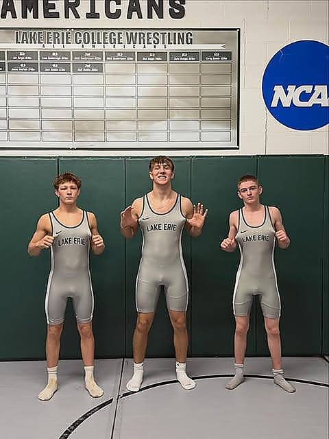 Three young men in matching grey college wrestling singlets pose together inside a wrestling room.