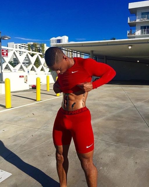 A muscular man in red sportswear poses outdoors, revealing his abs.