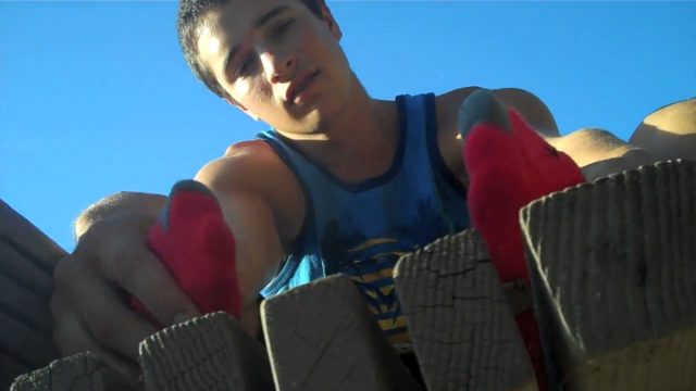 A young man in a blue tank top and red socks is seen from below, appearing to climb over a wooden fence outdoors.