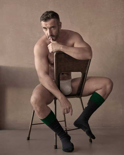 A muscular, bearded man poses sitting backwards on a metal chair wearing white ribbed underwear and tall green and black sports socks.