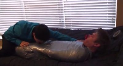 One man is tightly wrapped in plastic while another man leans over him on a couch.