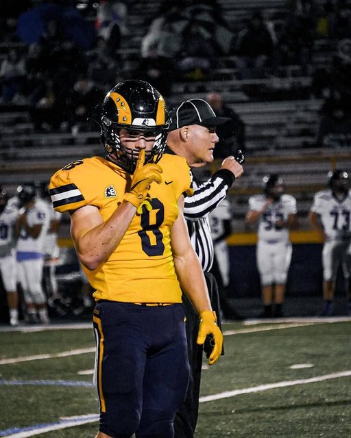 A muscular football player in full gear makes a shushing gesture while standing on a field next to a referee.