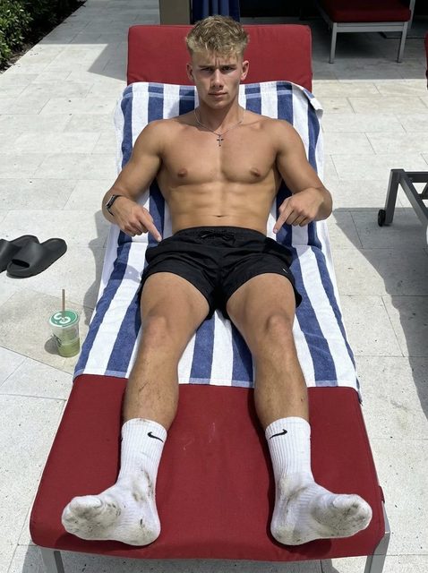 A shirtless, muscular young man in black gym shorts and soiled white crew socks reclines on a poolside lounge chair.