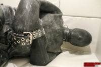 A person completely encased in a heavy black rubber suit and hood lies bound with thick leather grommet straps.