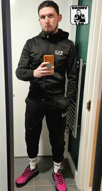 A man in a black tracksuit, white socks, and pink sneakers takes a mirror selfie indoors.