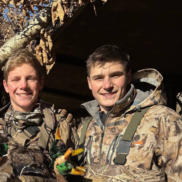 Two young men in camouflage hunting gear smile while holding ducks outdoors.