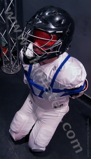 A person in a full lacrosse uniform and helmet is kneeling while restrained with blue rope.
