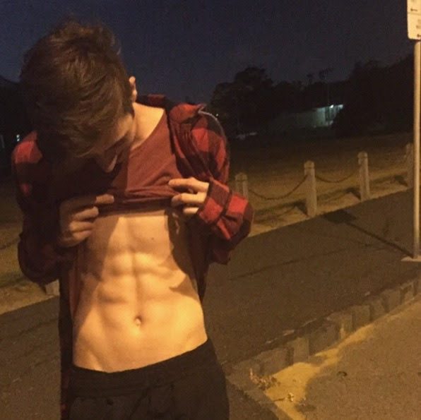 A young man in a plaid shirt reveals his muscular abs outdoors at night.