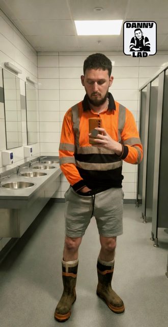 A man in a high-visibility shirt, shorts, and boots takes a selfie in a public restroom.