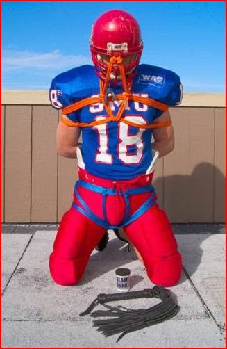 A man in a football uniform is restrained with ropes and kneels outdoors next to a flogger.