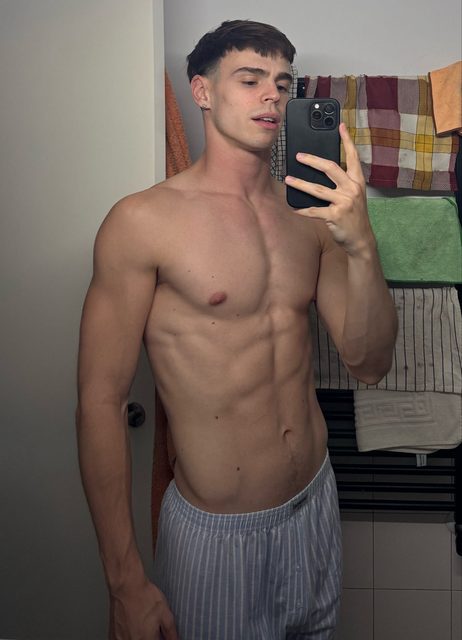 A young, muscular man takes a mirror selfie in a bathroom.