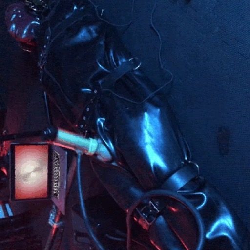 A person in a black latex suit, hood, gloves, and harness is restrained, with red light illuminating the scene.