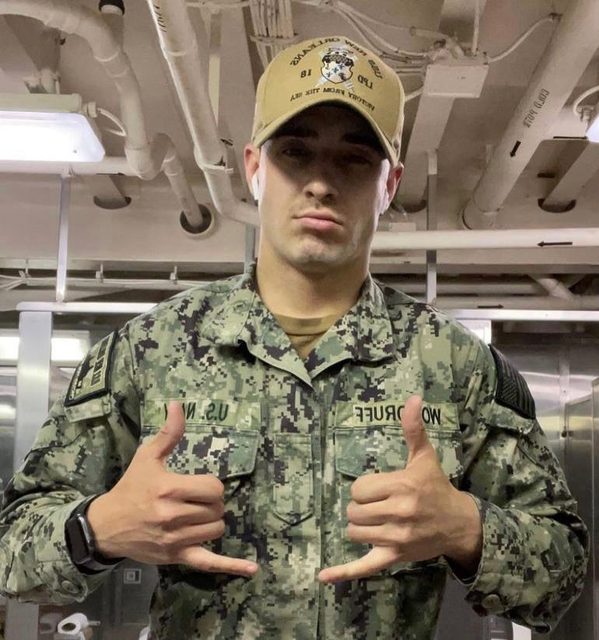 A man in a military camouflage uniform and cap poses with hand gestures indoors.