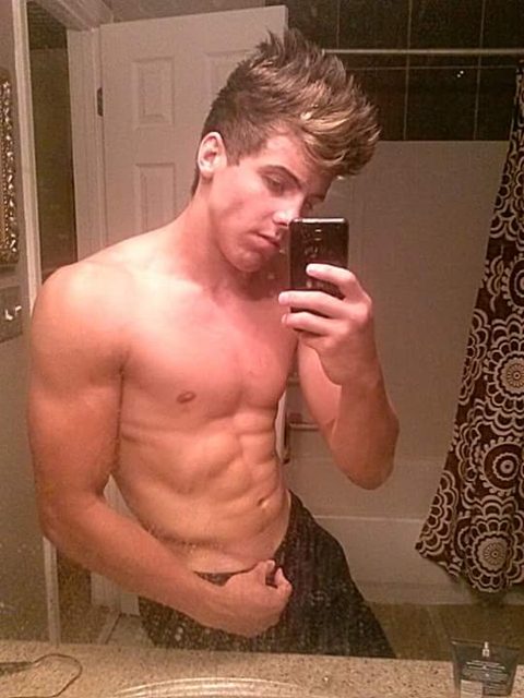 A muscular man takes a mirror selfie in a bathroom.