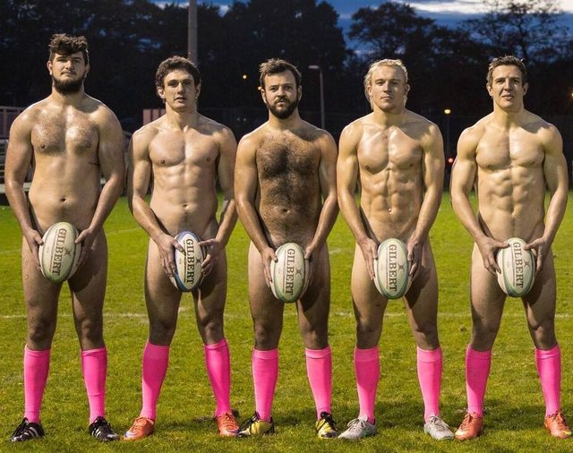 Five shirtless men pose on a grassy field, holding rugby balls over their crotches and wearing pink athletic socks.