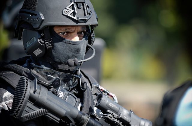 A person in a tactical military uniform with a helmet and balaclava holds a rifle outdoors.