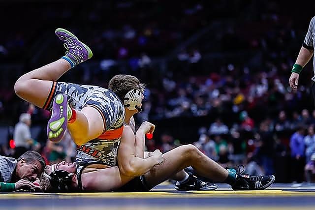 Two wrestlers in singlets and headgear compete on a mat inside a crowded sports arena.