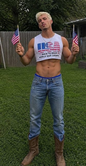 A muscular man with bleached blonde hair poses outdoors in a cropped USA tank top, denim jeans, and cowboy boots while holding American flags.