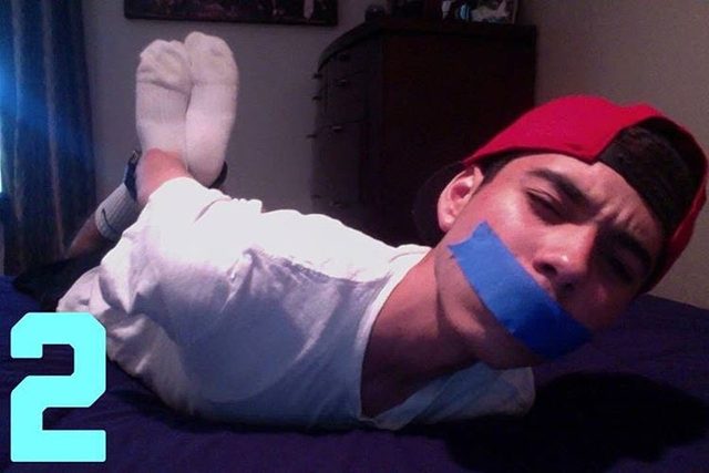 A young man with a red cap is lying prone on a bed, gagged with blue tape and with his feet restrained.