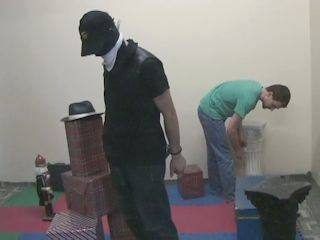 A man in a dark mask and vest stands indoors near another man bending over.