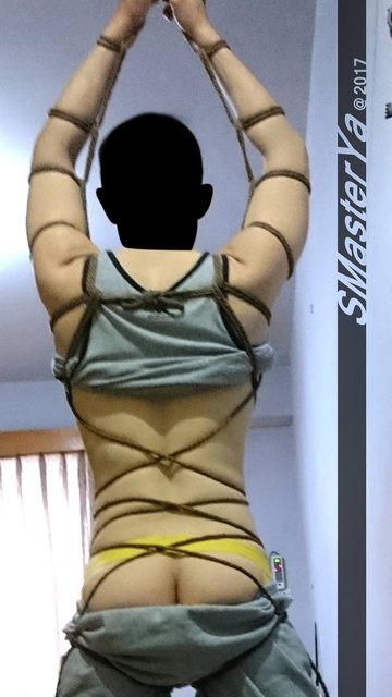 A man in rope bondage with yellow underwear and grey sweatpants is restrained in a bedroom.