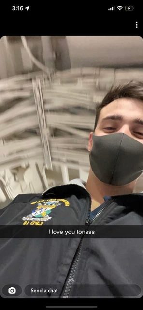 A young man takes a selfie indoors wearing a jacket and a face mask.