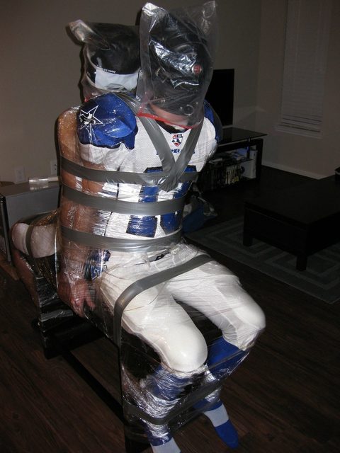 A person in a full American football kit and leather hood is heavily bound with plastic wrap and duct tape, including a plastic bag over their head.