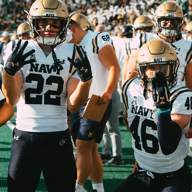 Two muscular football players pose in full Navy uniforms, gold helmets, and athletic gloves on an outdoor stadium field.