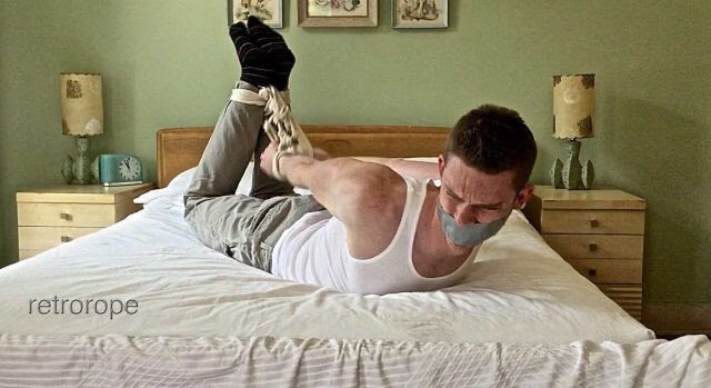 A man is hogtied on a bed with his mouth taped shut.