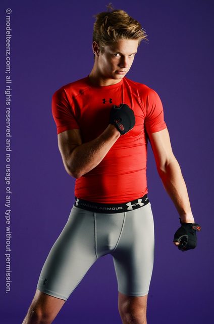 An athletic young man poses in a red compression shirt, grey compression shorts, and workout gloves against a purple background.