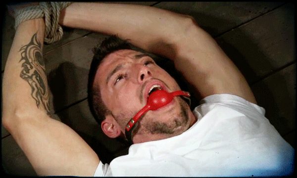 A muscular man with tattoos is tied up with rope and gagged.