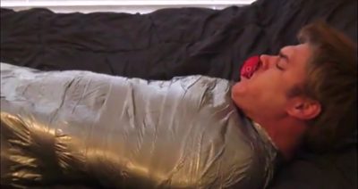 A man is restrained in silver material with a red gag in his mouth while lying on a dark surface.