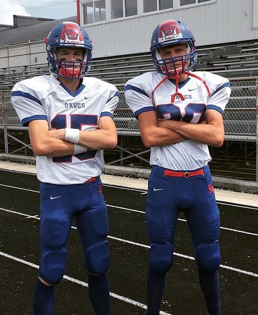 Two athletic young men in full football gear and helmets pose with arms crossed on an outdoor track.