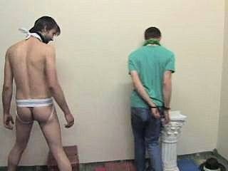 Two men are in a room, one is shirtless in a jockstrap and harness, the other is wearing a t-shirt and jeans with his hands cuffed behind his back.